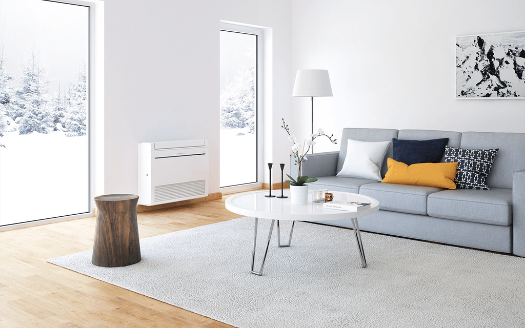 Ett modernt vardagsrum med en vit Mitsubishi Electric golvmodell värmepump monterad vid väggen. Värmepumpen håller rummet varmt under vintern, vilket syns genom de snötäckta träden utanför fönstret.