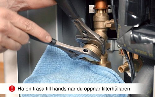 En närbild av händer som använder två skiftnycklar för att öppna filterhållaren på en värmepump. En blå trasa används för att fånga upp eventuellt vatten under filterrengöringen. Regelbunden rengöring av filtret säkerställer optimal drift och förhindrar att smuts blockerar vattenflödet i systemet.