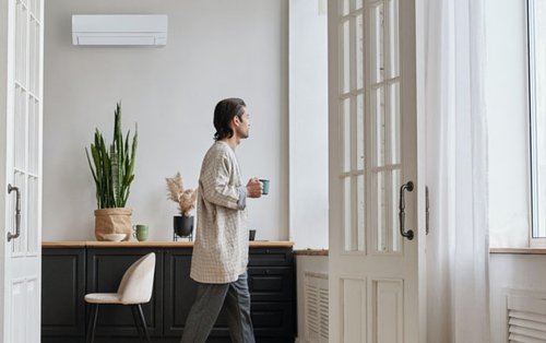 En person går genom ett ljust rum med en kopp kaffe. På väggen ovanför finns en vit Mitsubishi Electric luftkonditionering som bidrar till en behaglig inomhustemperatur.